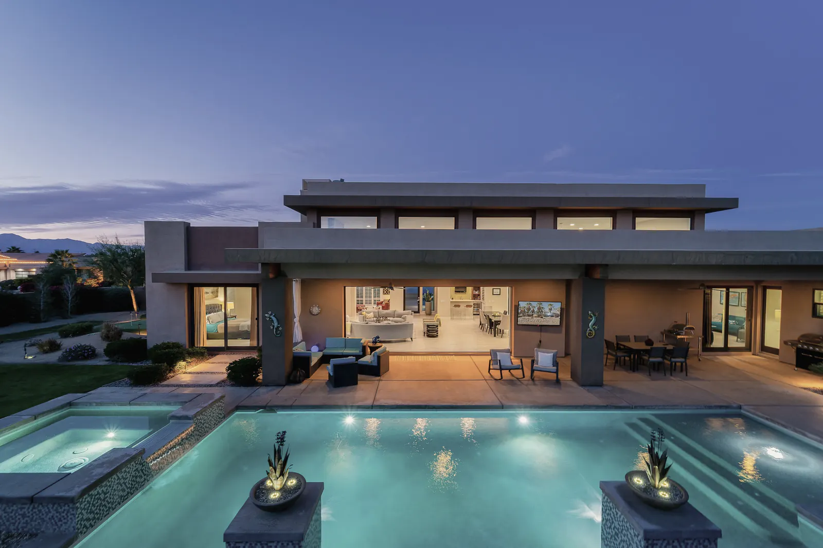 A desert home wrapped around a pool, lit from inside at dusk