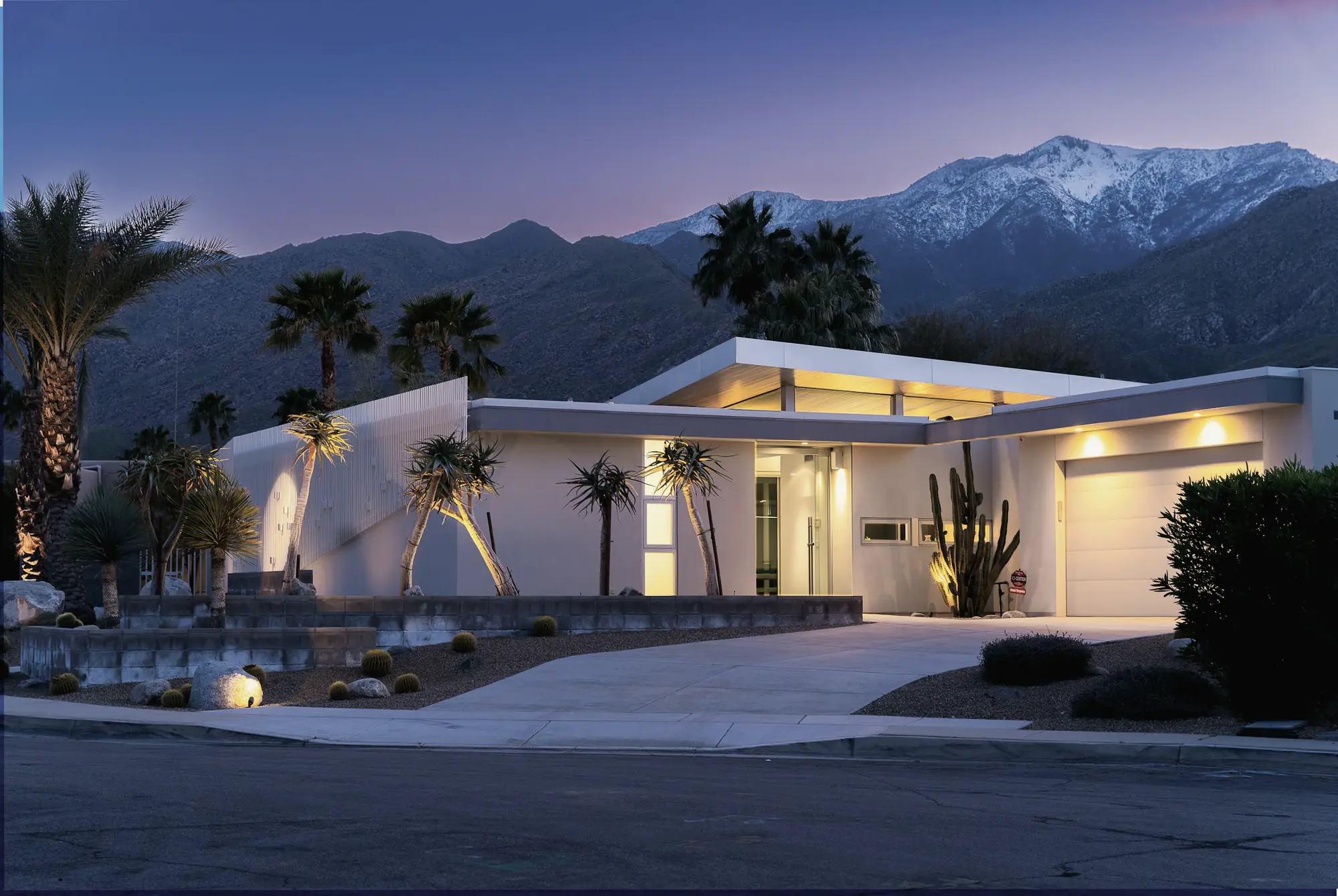 A low desert-modern home lit up at dusk below the mountains